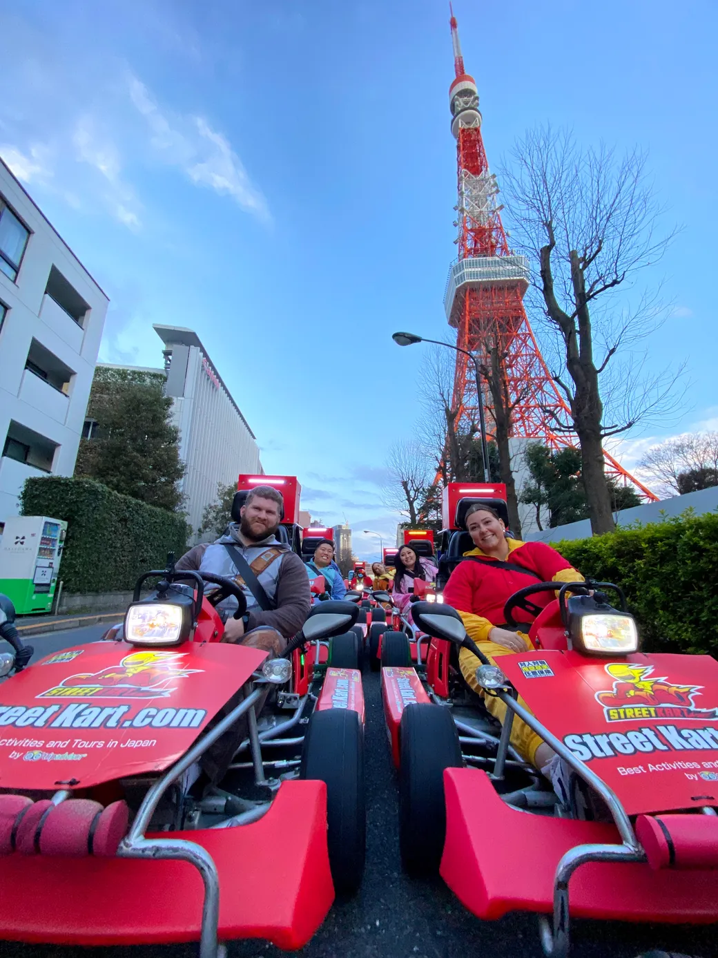 People ride red street-kart bikes on a city street with Tokyo Tower in the background, sunlight-blue sky above.