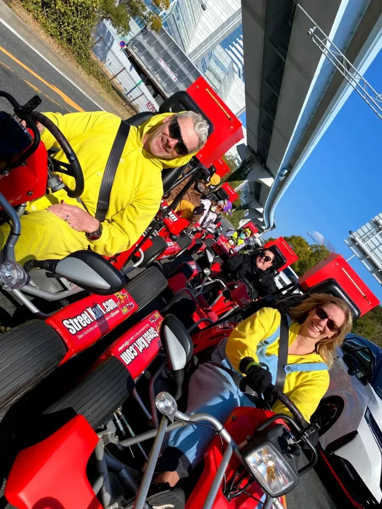 A group of people smiling and riding bright red pedal-powered tour trikes in a line along an urban street on a sunny day, wearing helmets and sunglasses.