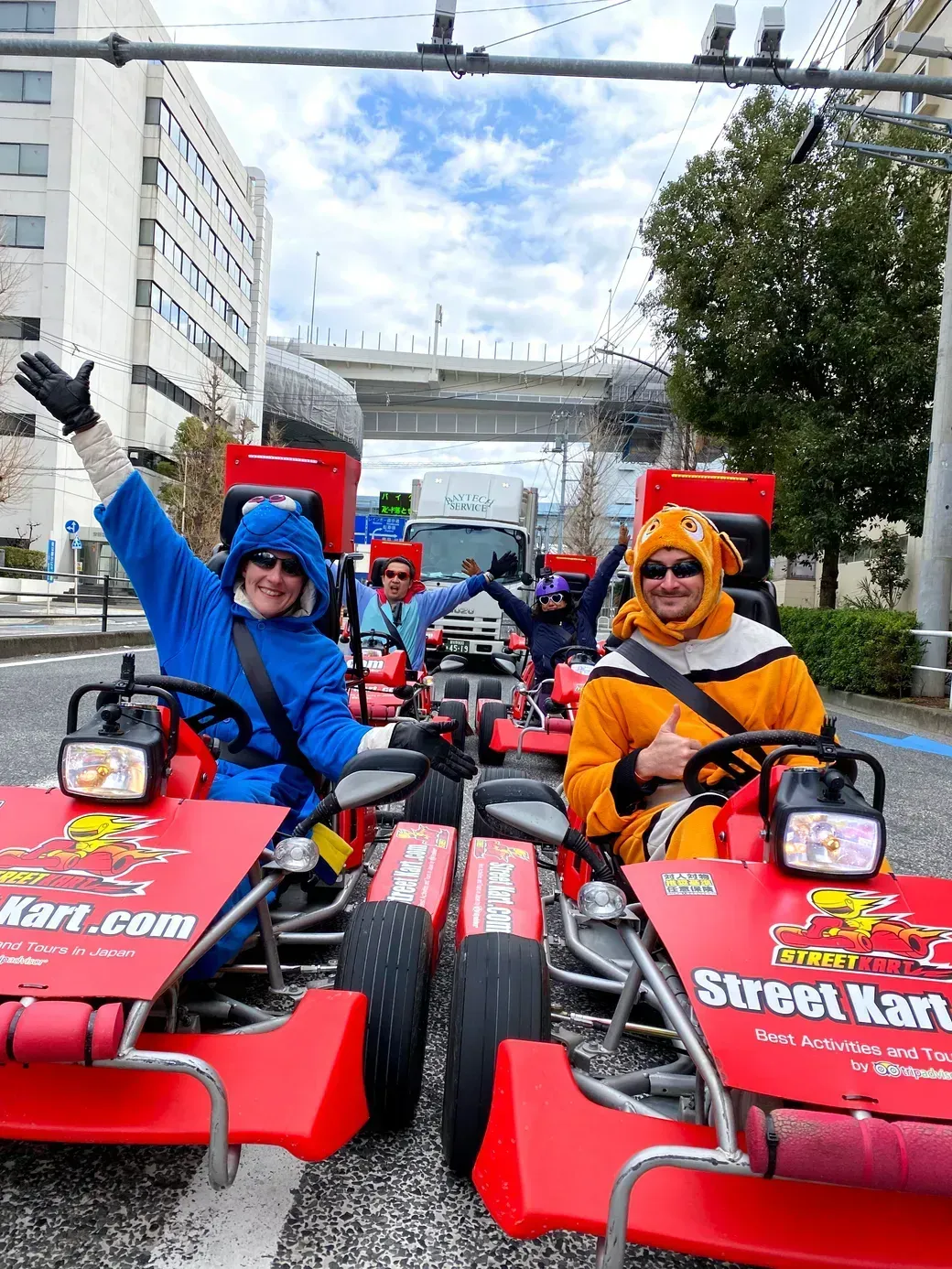 Group of people in red go-karts wearing colorful animal costumes, waving at the camera on an urban street ride.