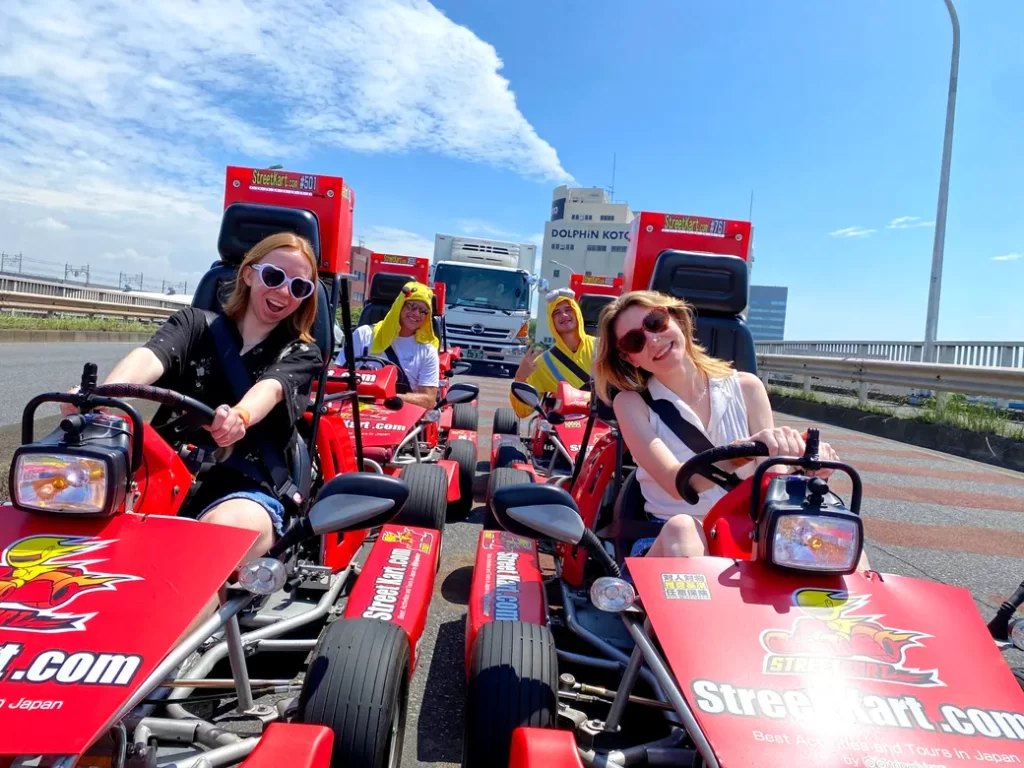 Group of friends in red go-karts racing on a highway, smiling and posing for the camera with trucks in the background on a sunny day.